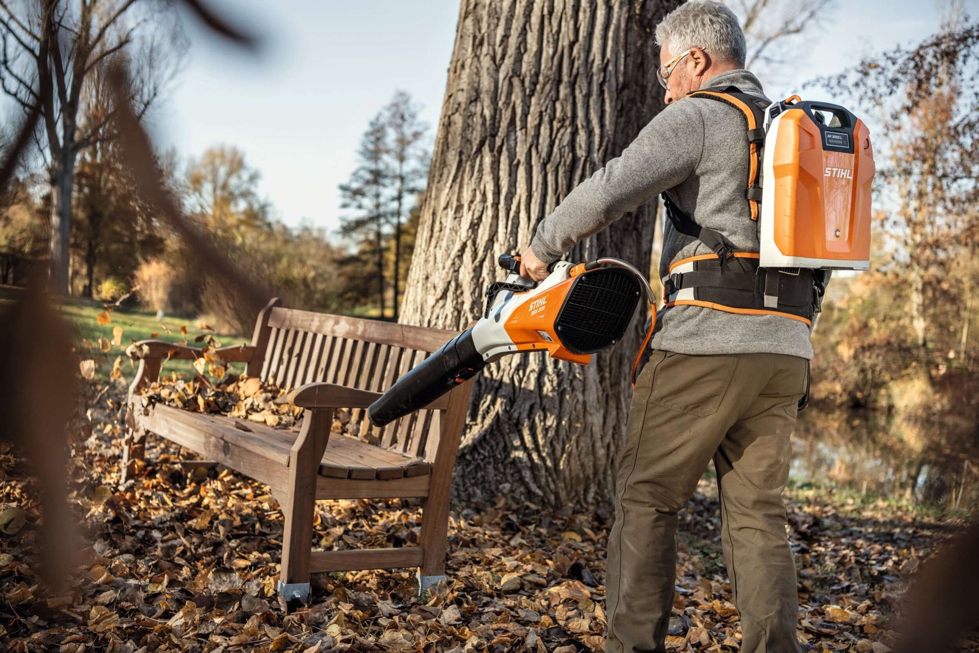 Tree surgeon using STIHL blower vacuum accessories to clear leaves near a park bench in autumn.