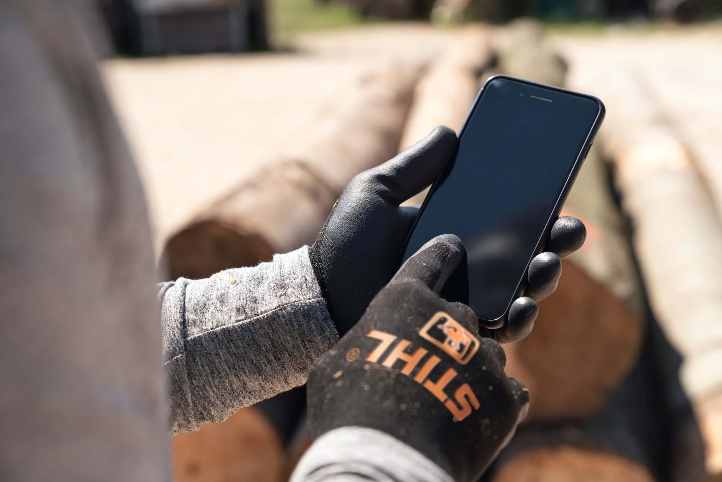 Tree surgeon wearing protective gloves while using a smartphone on a job site with logs in the background.