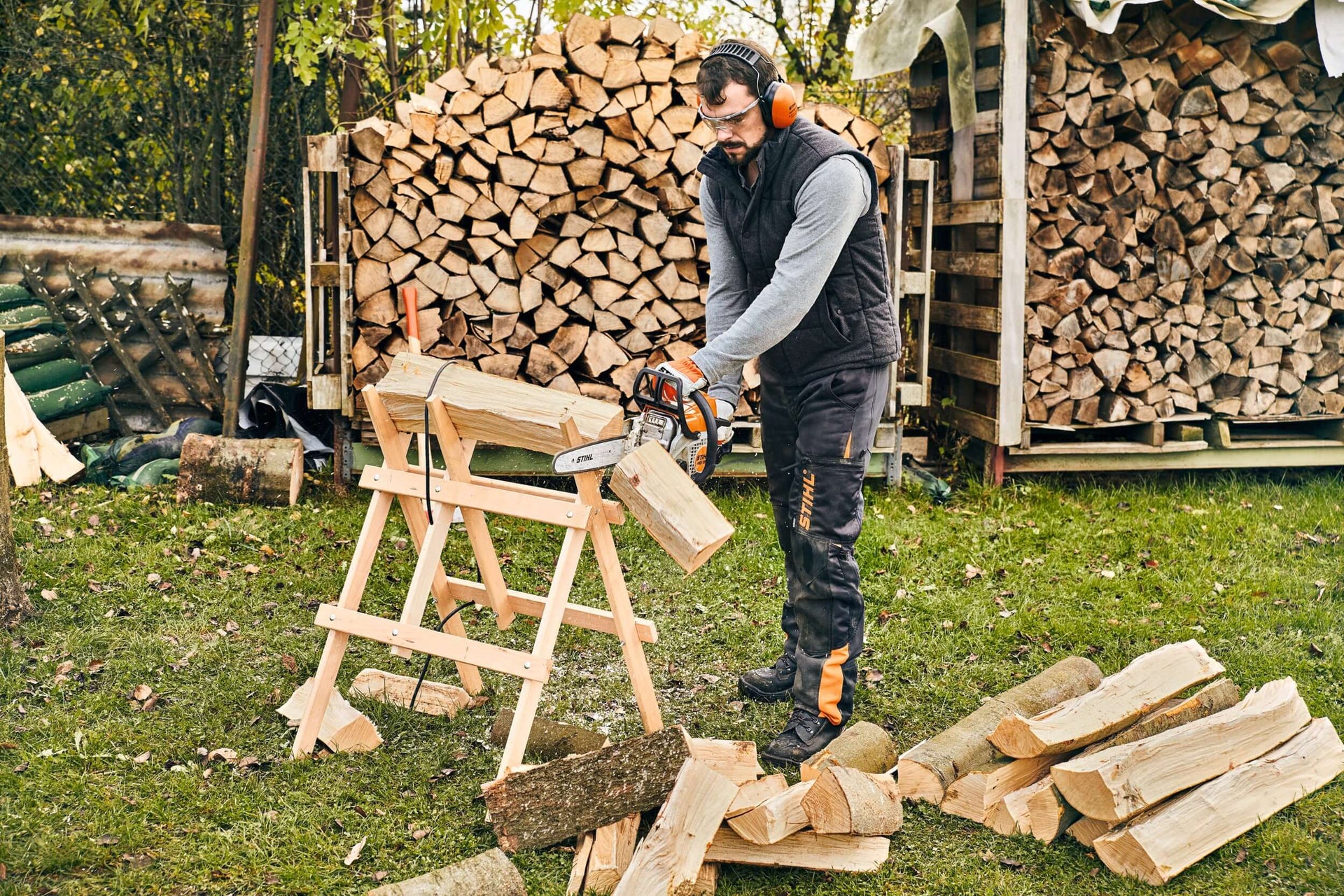 Man using a chainsaw on a wooden sawhorse to cut logs in a yard, showcasing essential tools for tree surgeons.