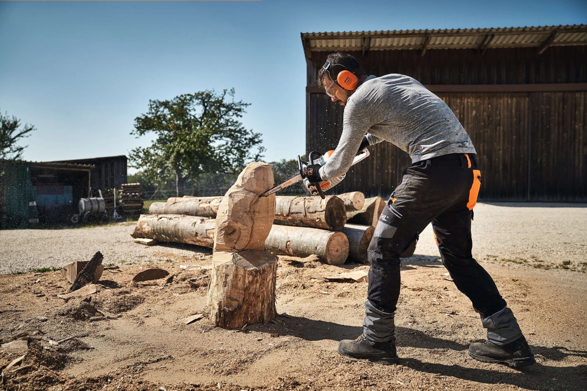 A tree surgeon using a STIHL chainsaw to carve wood outdoors, surrounded by logs and natural scenery.