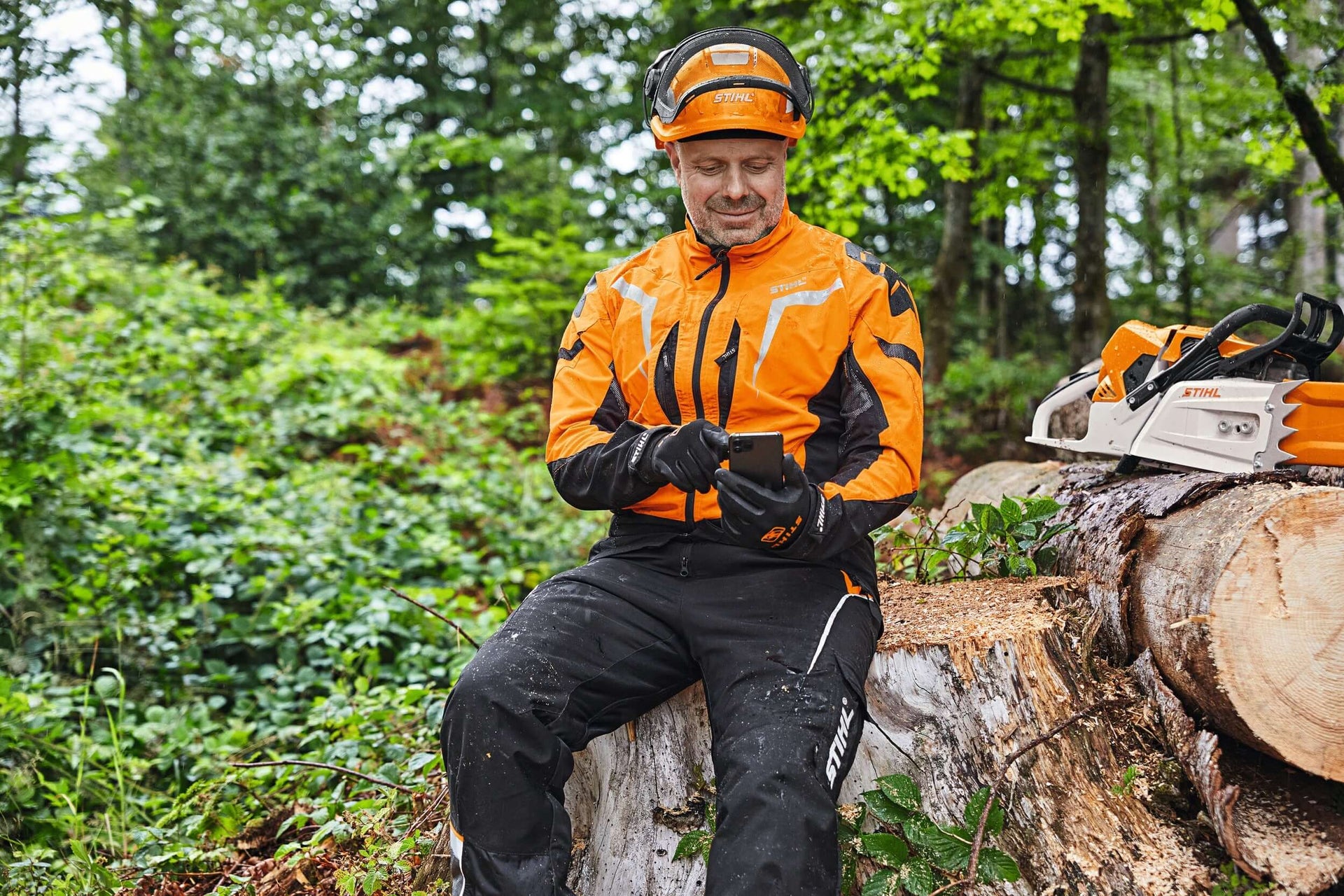 Tree surgeon in STIHL chainsaw trousers and helmet checking equipment in a forested area.