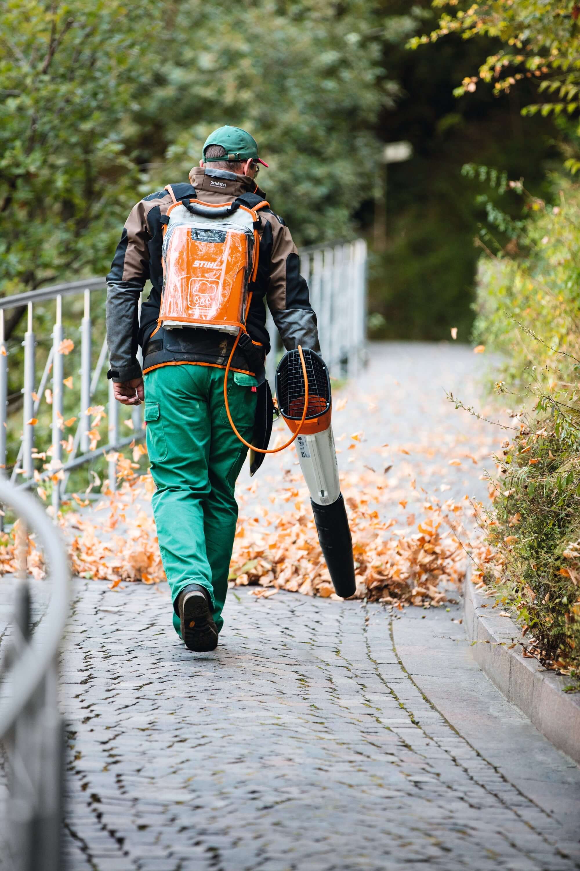 Tree surgeon using a STIHL backpack leaf blower to clear leaves from a stone pathway in a wooded area.