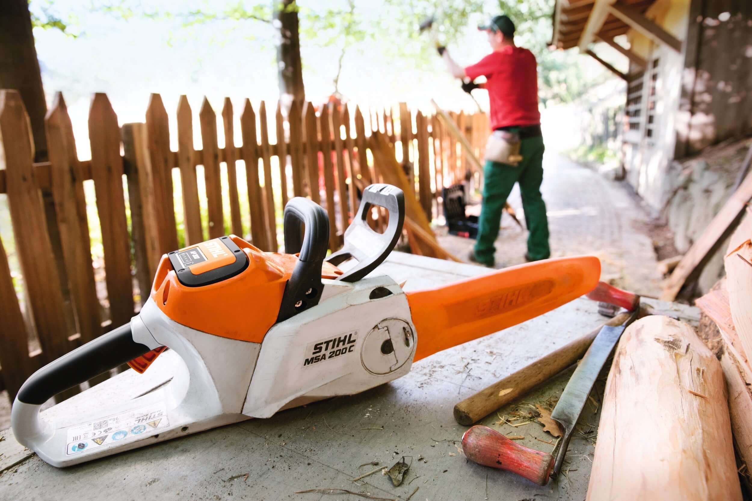 STIHL battery chainsaw on a workbench with a tree surgeon in the background performing outdoor tasks.