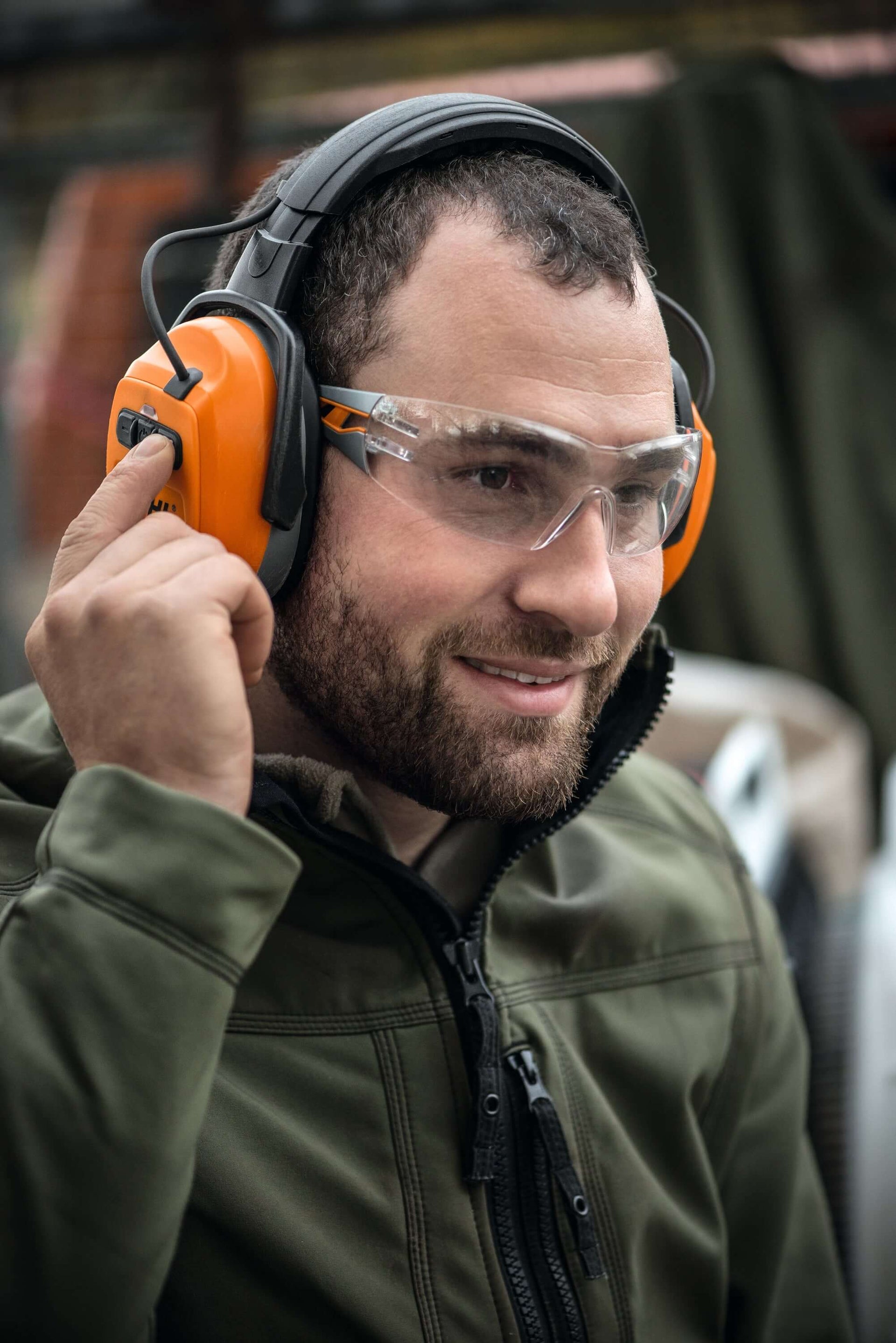 Tree surgeon adjusting Bluetooth ear defenders with protective goggles in a work environment.