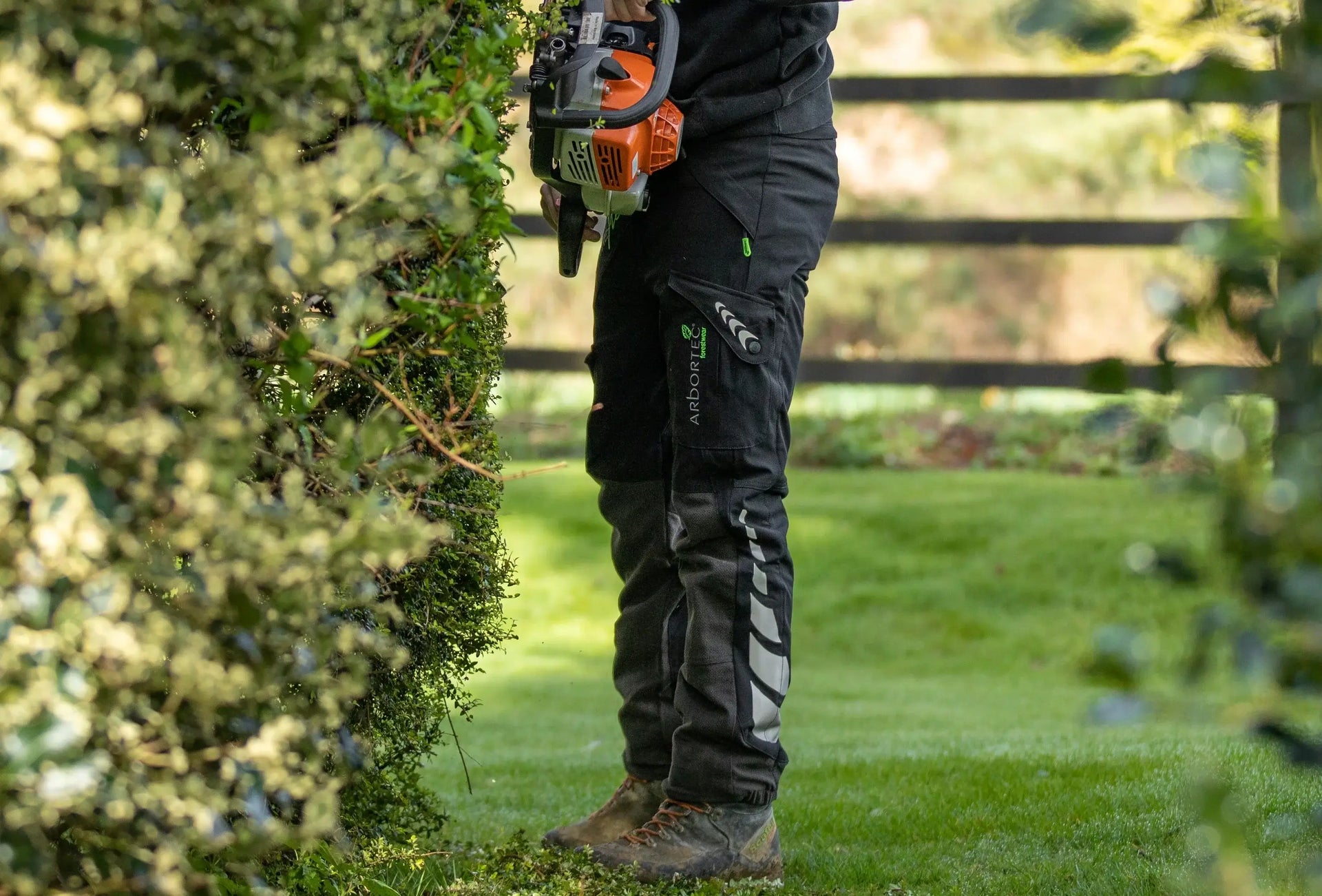 Arbortec Type A chainsaw trousers worn by a tree surgeon while trimming hedges in a garden.