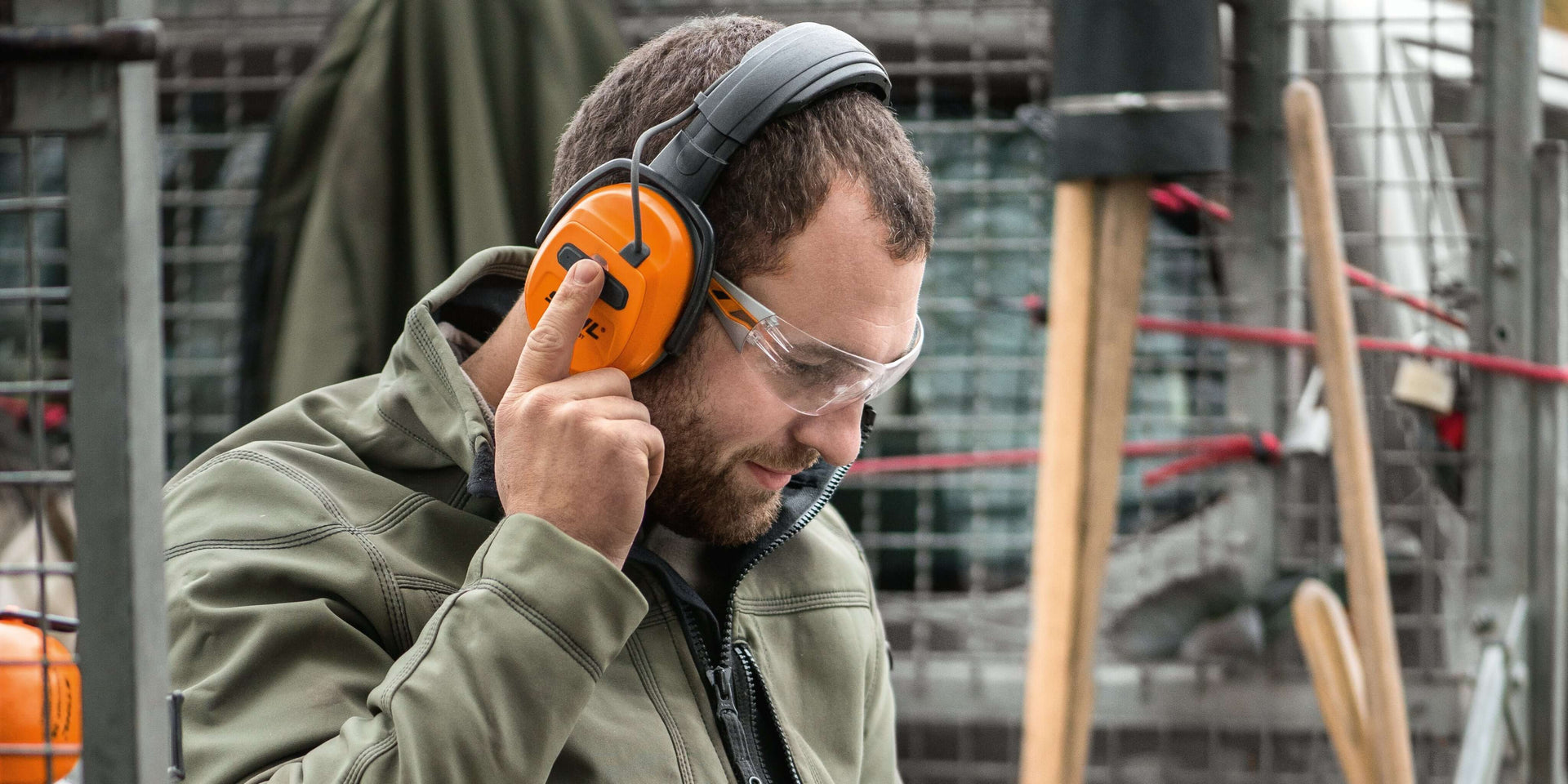 Tree surgeon wearing Bluetooth ear defenders and safety goggles, adjusting his hearing protection on a worksite.