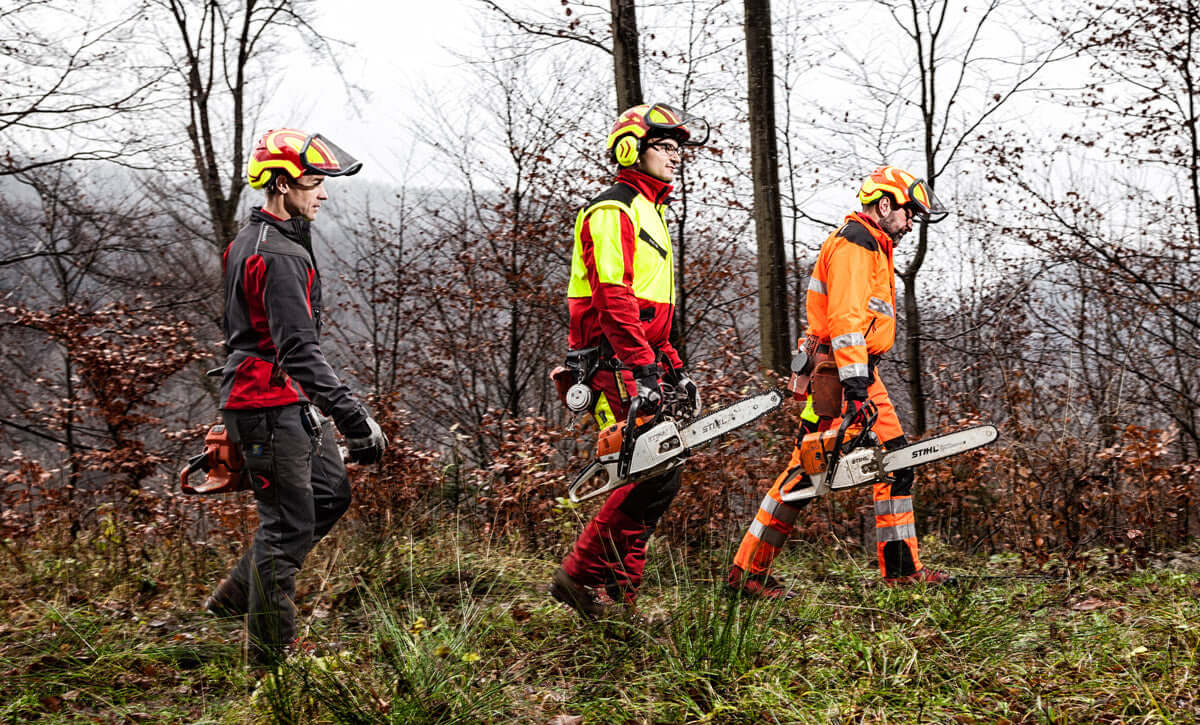 Three tree surgeons in high-visibility gear walking through a forest with chainsaws in hand, showcasing Pfanner workwear.