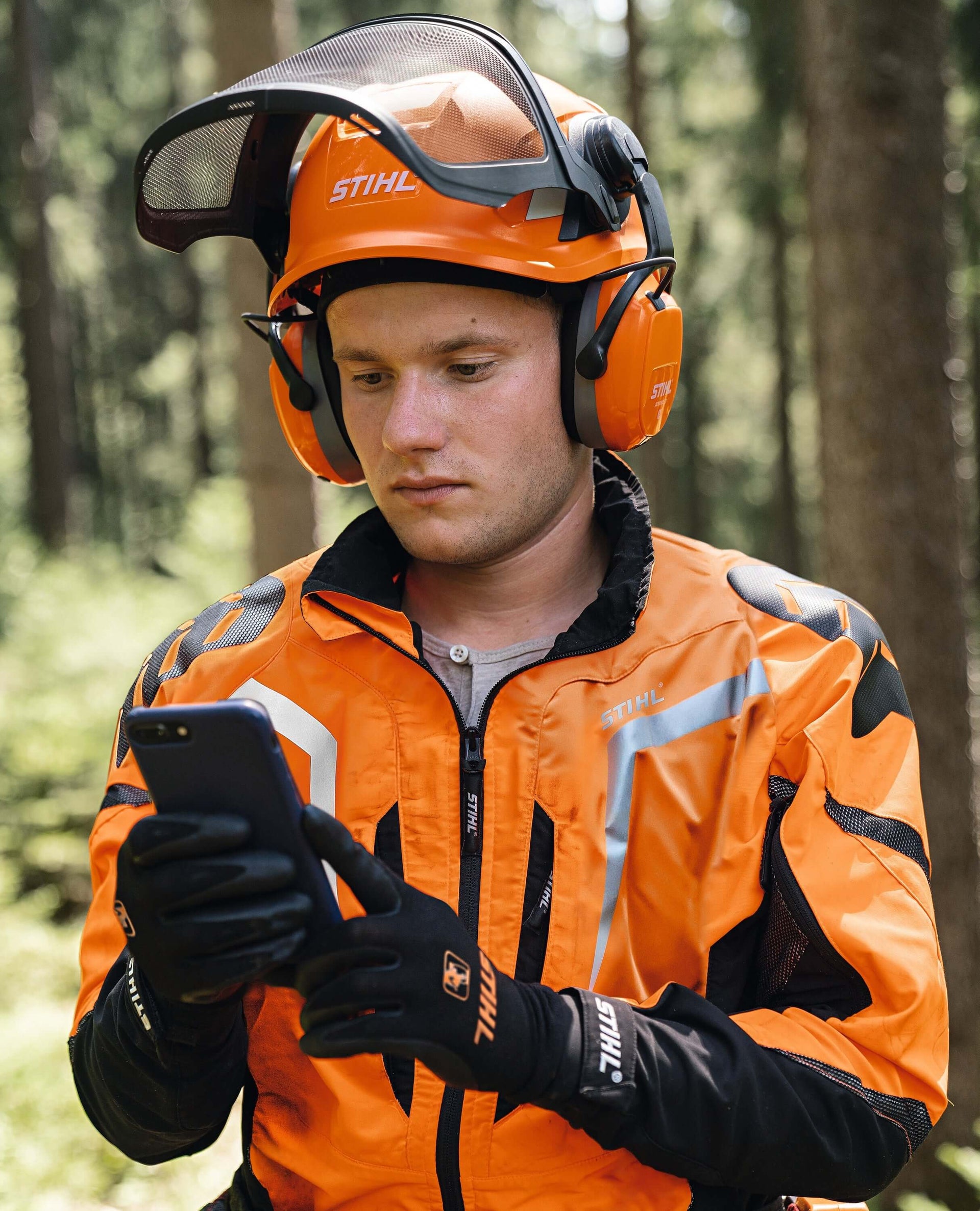 Tree surgeon in STIHL chainsaw helmet and protective gear, checking smartphone in a forest.