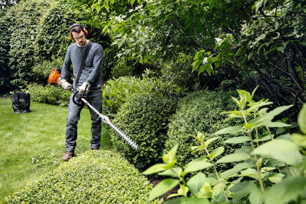 Tree surgeon using STIHL Kombi system to trim hedges in a lush garden setting.