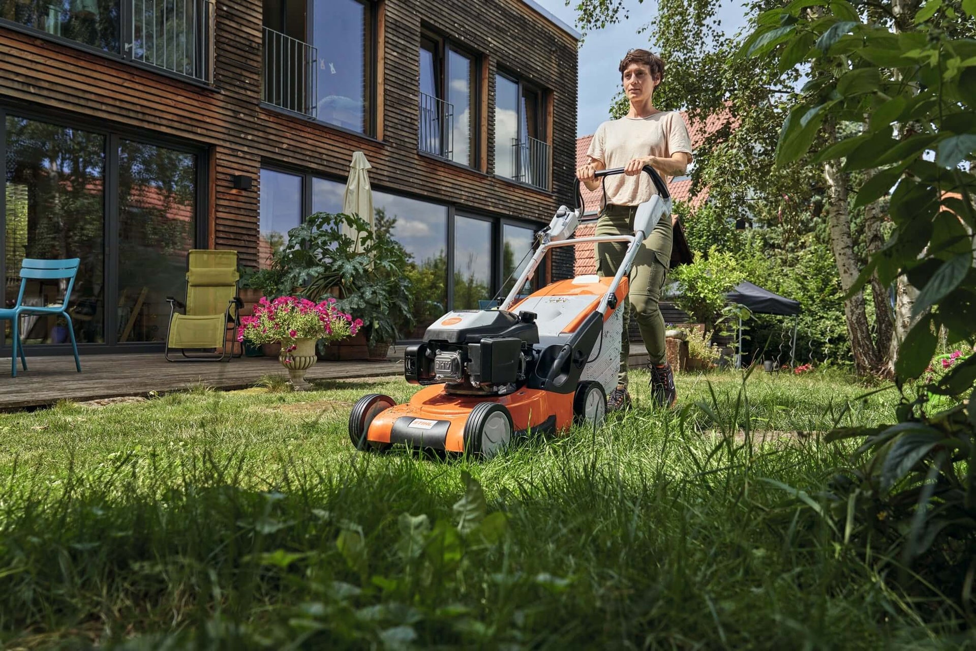 Person using a STIHL walk-behind lawn mower on a lush green lawn beside a modern home.