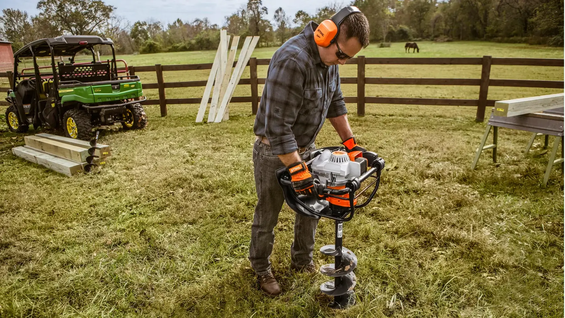 Man using a STIHL earth auger on a farm, surrounded by wooden posts and a utility vehicle, enhancing drilling efficiency.
