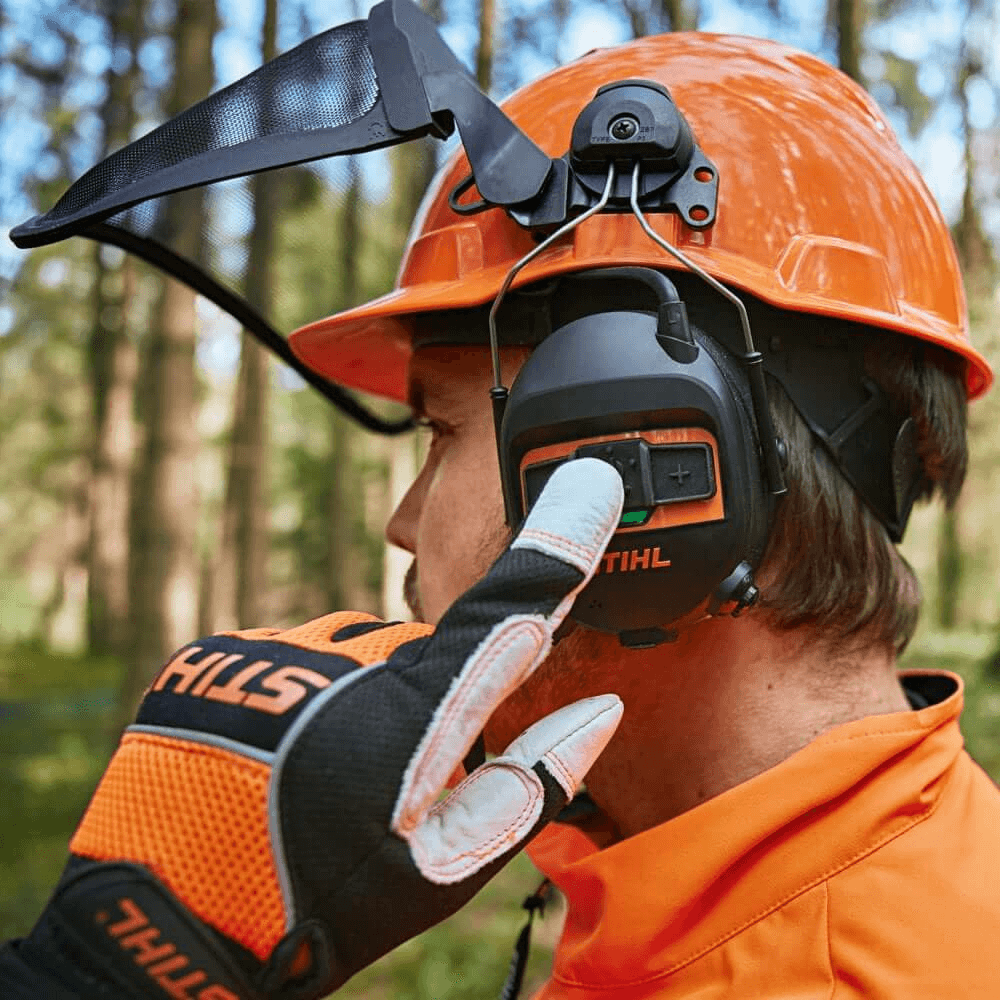 Tree surgeon wearing Bluetooth ear protectors while adjusting settings, combining safety and communication in a forest.