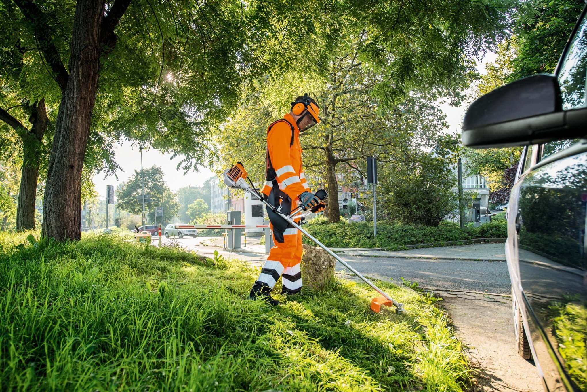 Tree surgeon in orange safety gear using a STIHL clearing saw to maintain a grassy area near a roadside.
