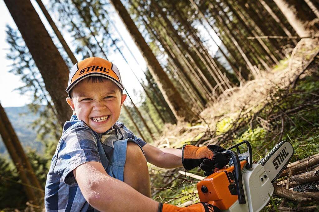 Young boy in a STIHL cap playing with a toy chainsaw, embodying the spirit of a tree surgeon in a forest setting.