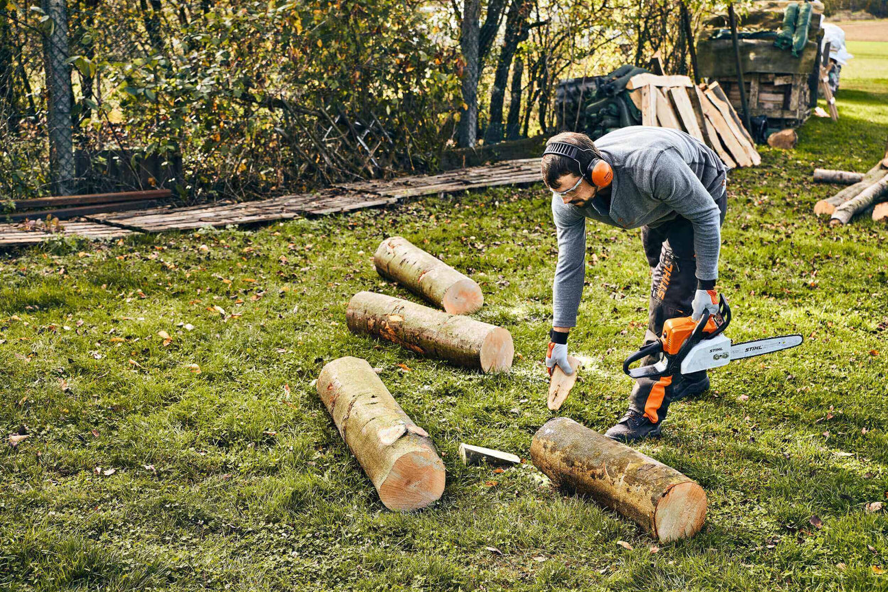 Person using a chainsaw to cut logs in a green field, wearing protective gear and focused on the task.