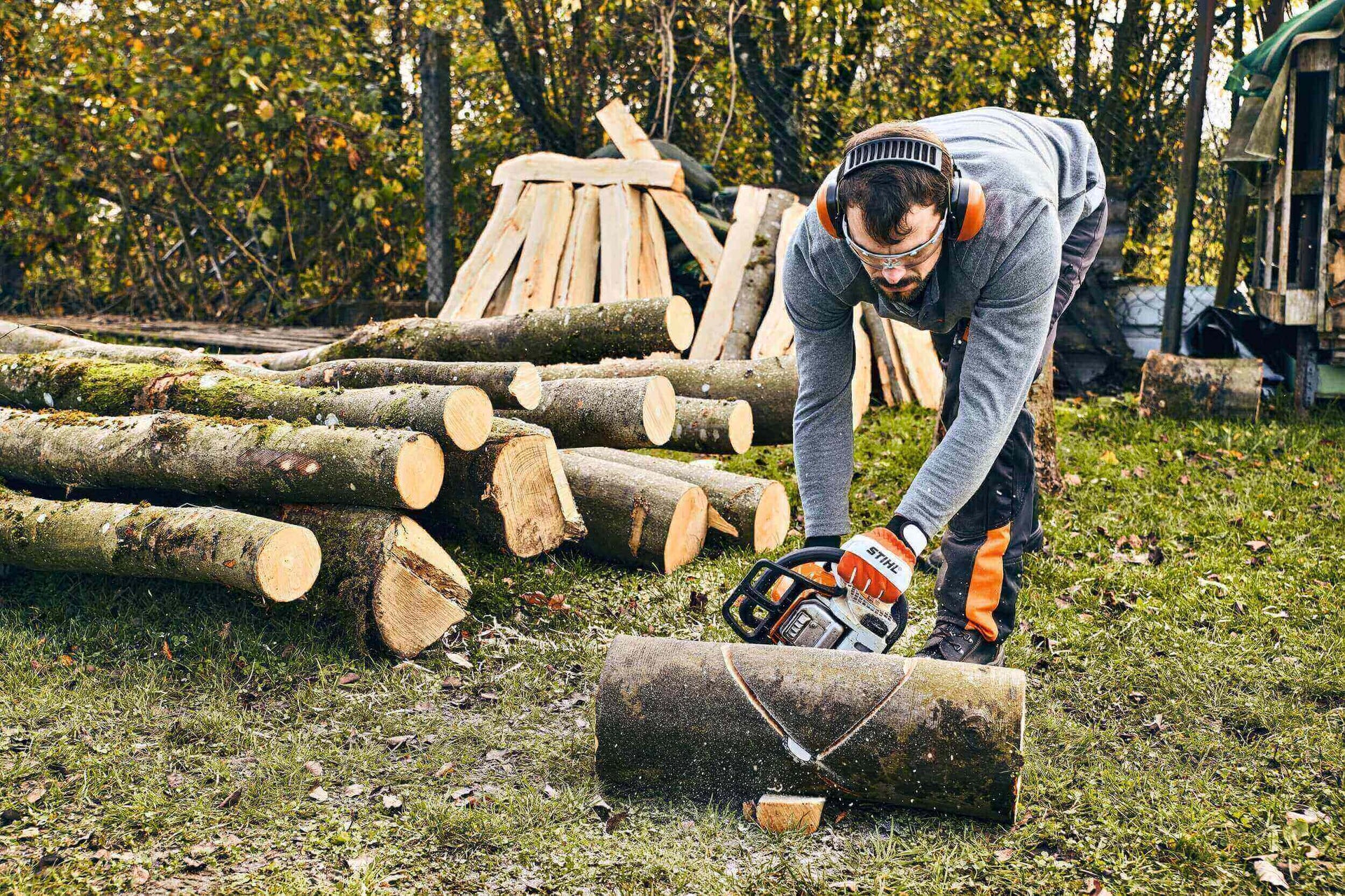 Tree surgeon using a chainsaw to cut logs in a wooded area with stacked firewood in the background.