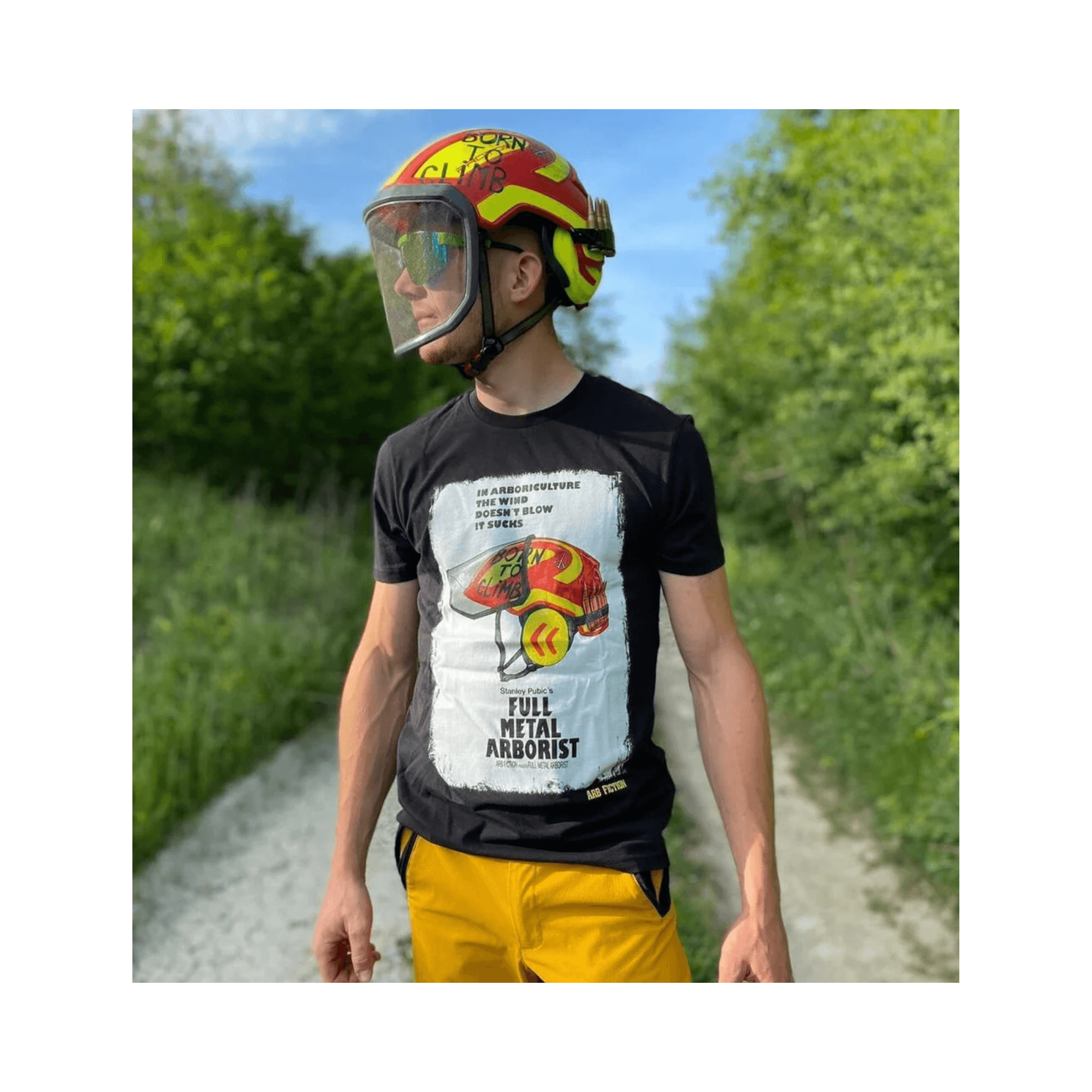 Person wearing a Full Metal Arborist T-Shirt and a helmet, standing on a path surrounded by trees.