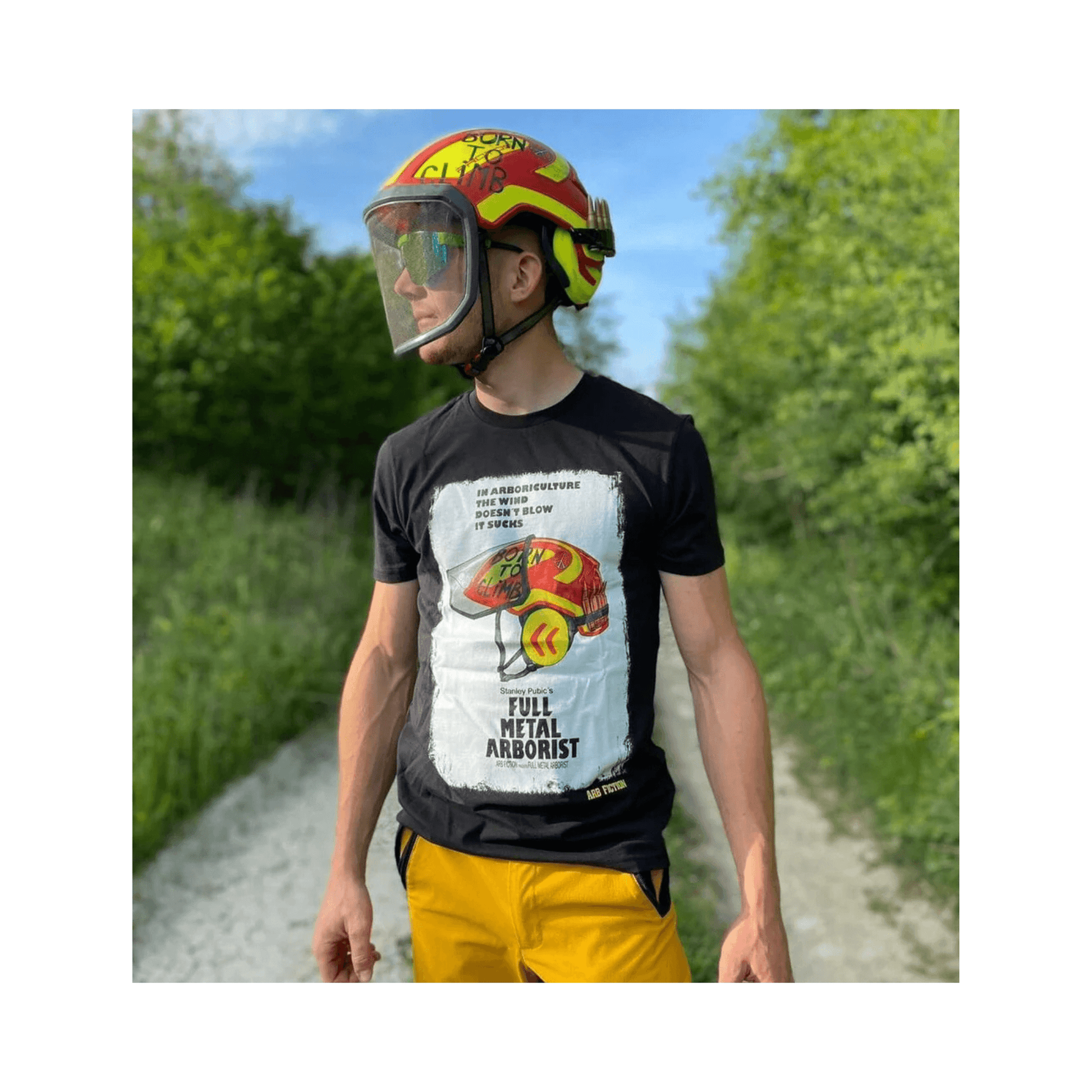 Person wearing a Full Metal Arborist T-Shirt and a helmet, standing on a path surrounded by trees.