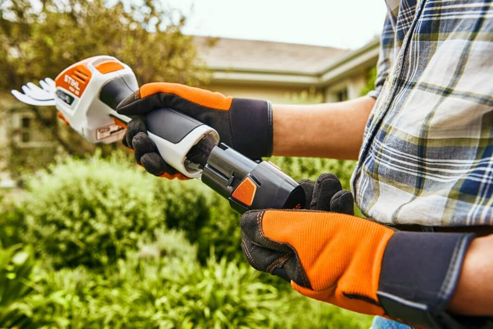 User holding STIHL HSA 26 cordless shrub shear, wearing gloves, ready for gardening tasks in a lush green garden.