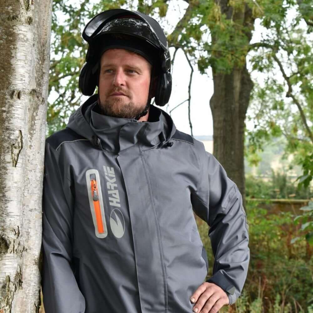 Man wearing Harkie Defiance Jacket Grey, designed for tree surgeons, standing against a tree with a helmet.
