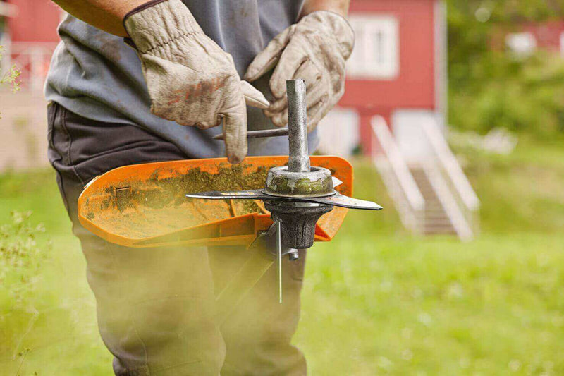 A person in gloves adjusts a string trimmer for gardening with a red building in the background.