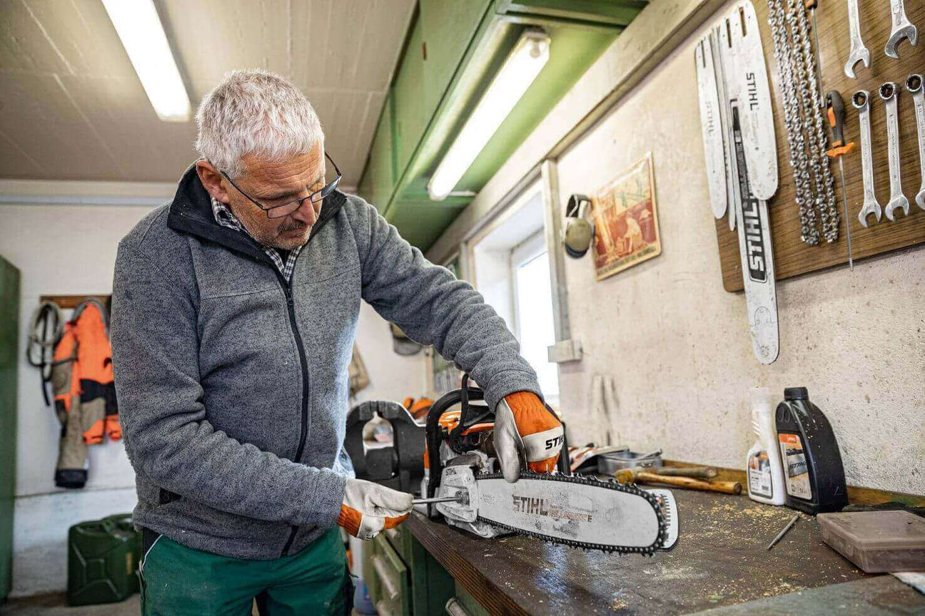 Man working on a chainsaw maintenance in a workshop, wearing gloves and glasses, with tools and STIHL branding.