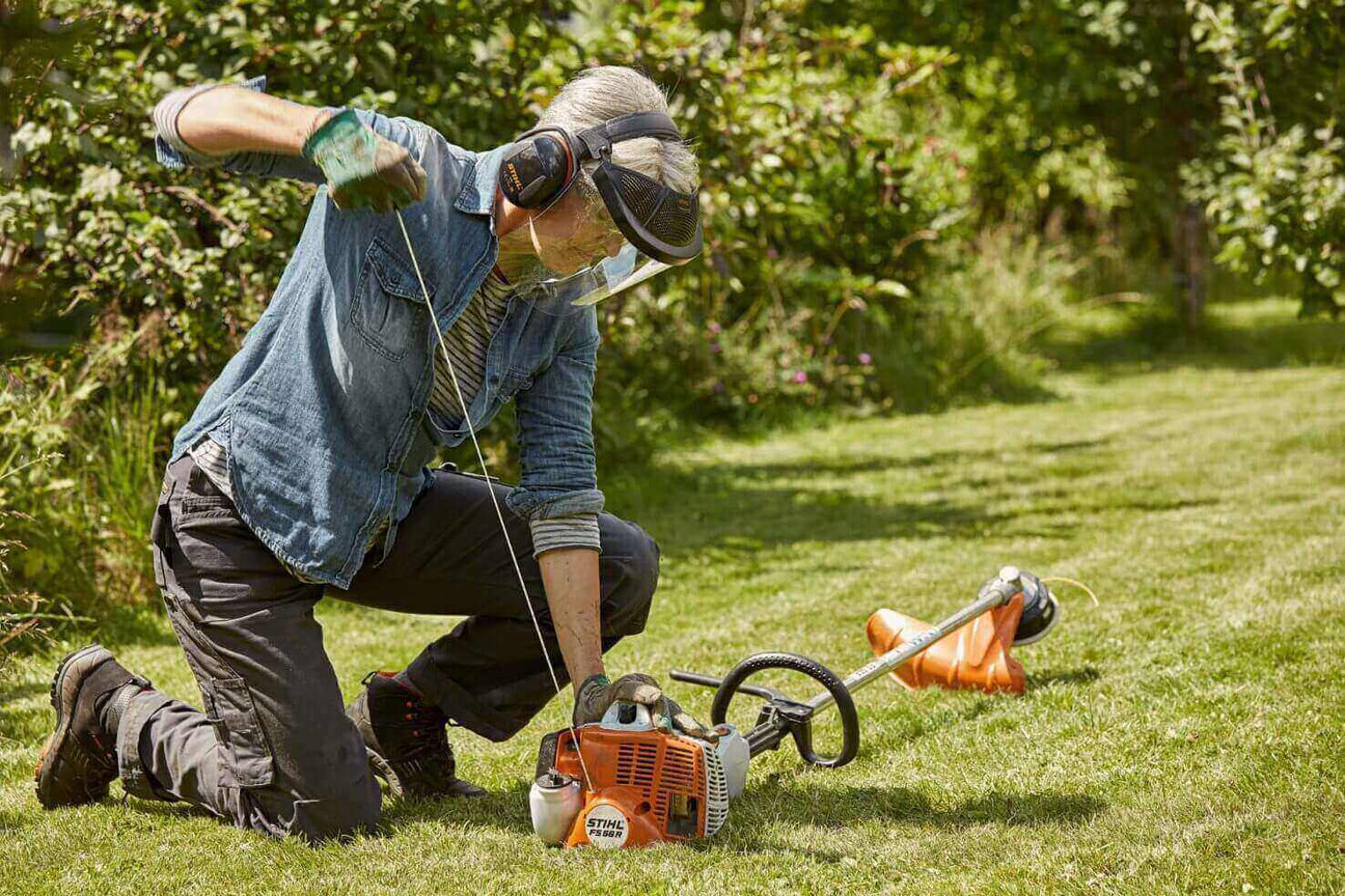 Person using a string trimmer in a garden, wearing protective gear and focused on maintaining the lawn.