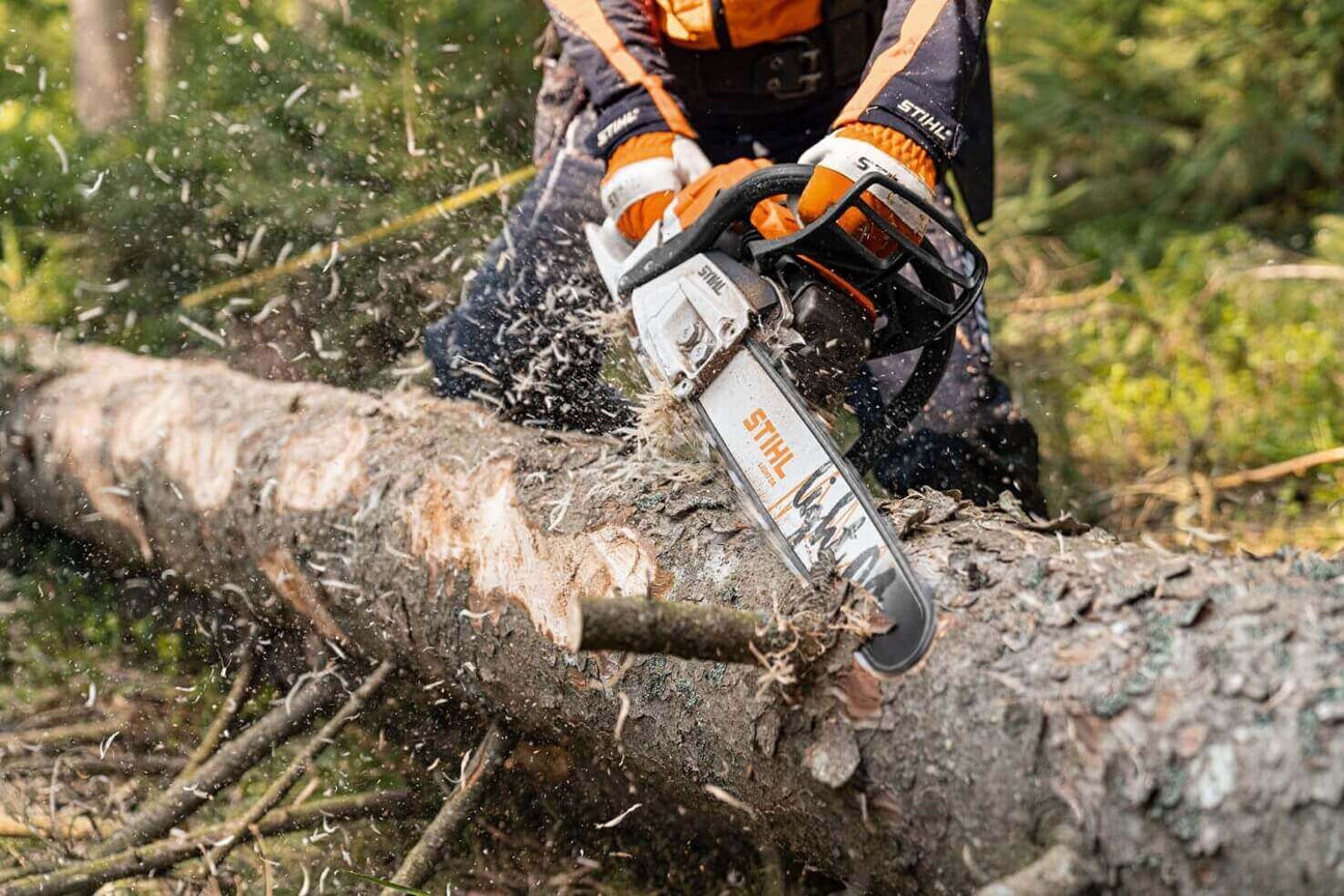 Person using a STIHL chainsaw to cut through a log, surrounded by flying wood chips and pine trees.