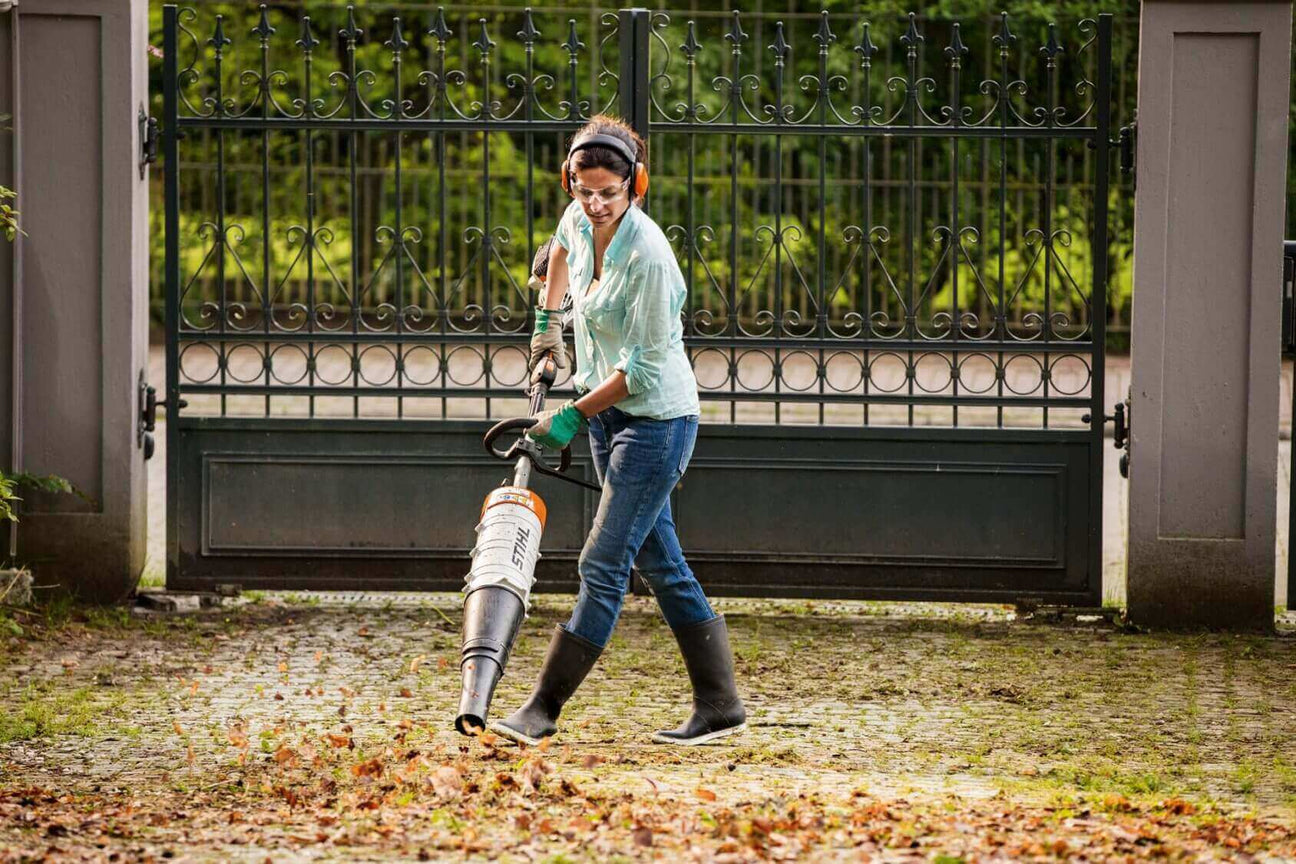 Woman using a leaf blower to clear leaves outside a gated entrance in a garden.