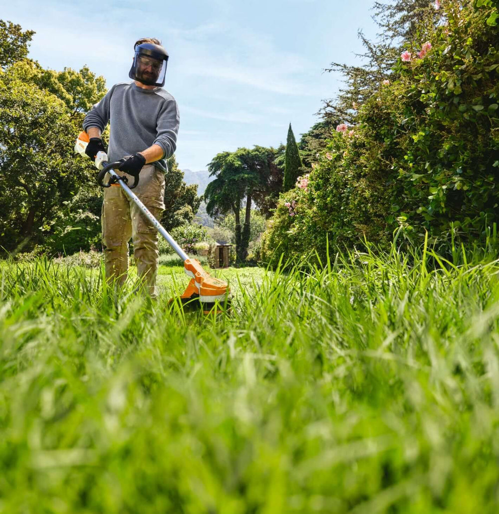 Man using STIHL FSA 70 R cordless strimmer in a lush green garden, wearing protective gear.