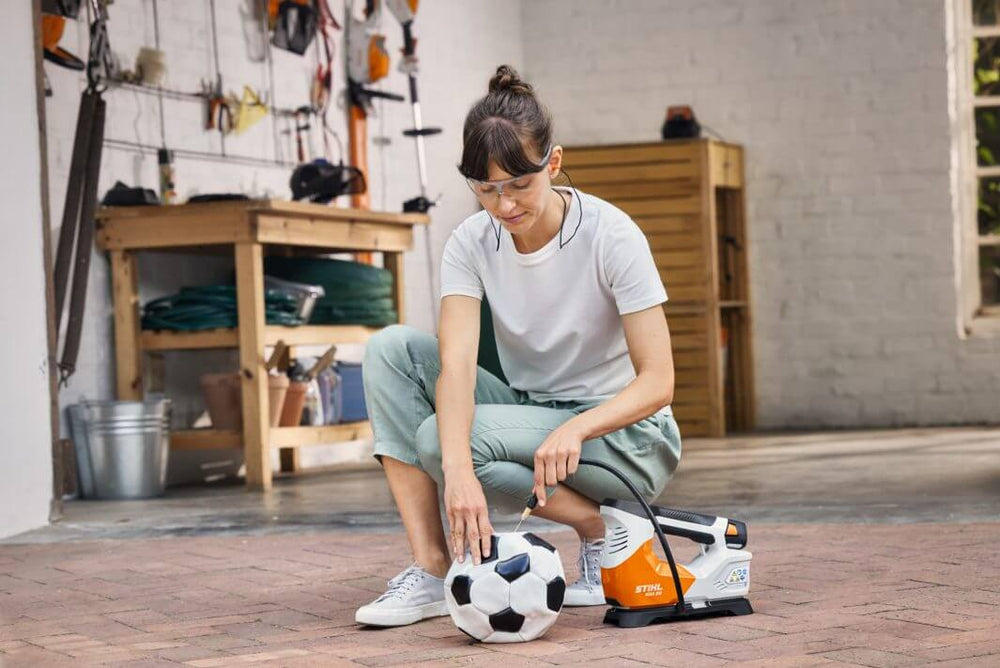 Woman using the STIHL KOA 20 cordless compressor to inflate a soccer ball in a garage, showcasing convenience for tree surgeons.