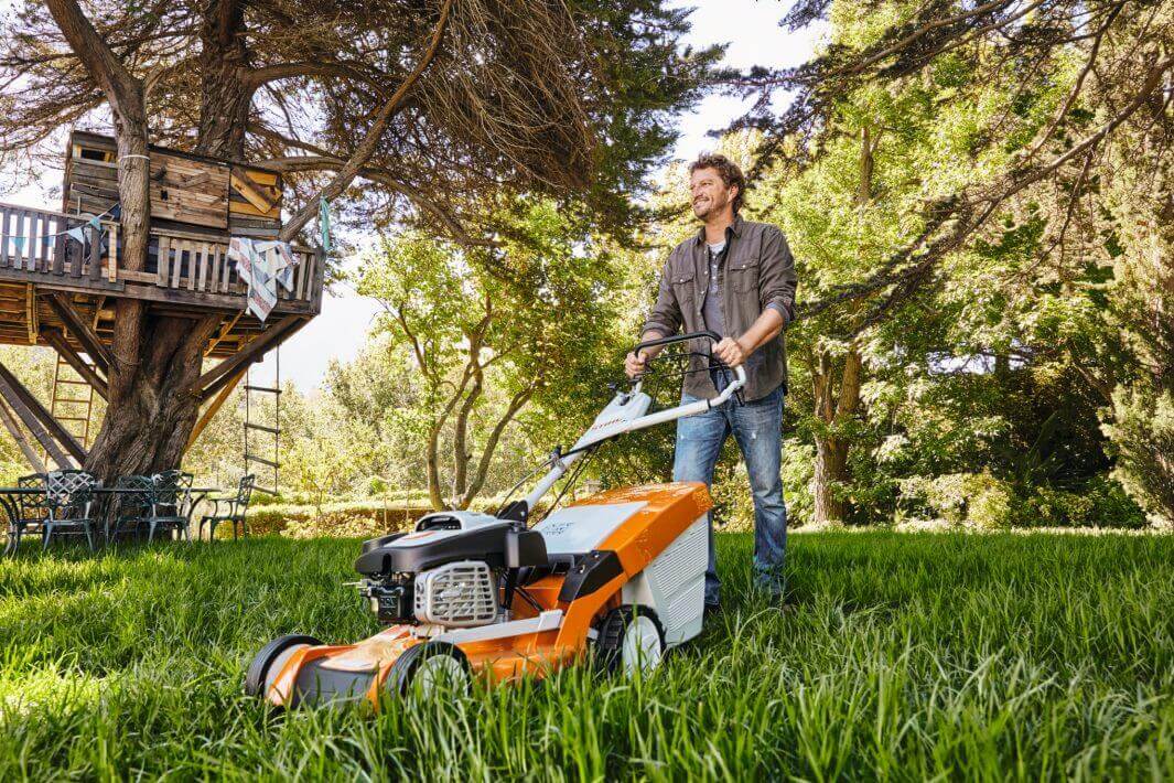 Man using STIHL RM 655 V petrol lawn mower in tall grass, surrounded by trees and a garden setup.