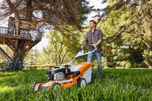 Man using STIHL RM 655 V petrol lawn mower in tall grass, surrounded by trees and a garden setup.