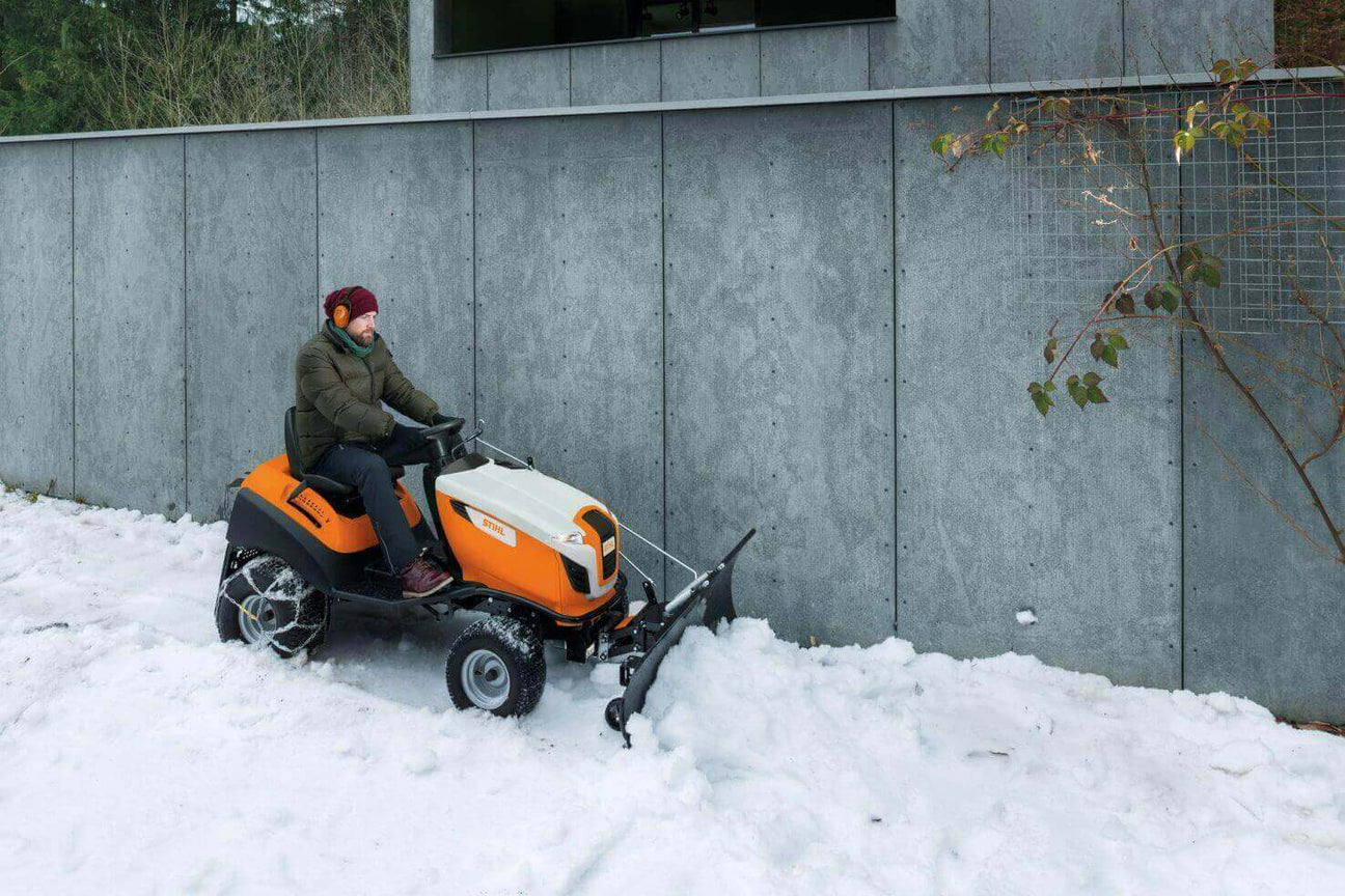 Person using a compact tractor to clear snow in a winter setting near a modern concrete wall.