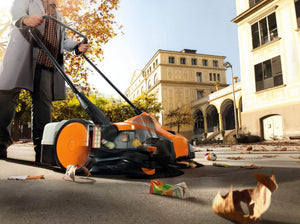 Person using STIHL KGA 770 cordless sweeping machine to clean debris from a city sidewalk.