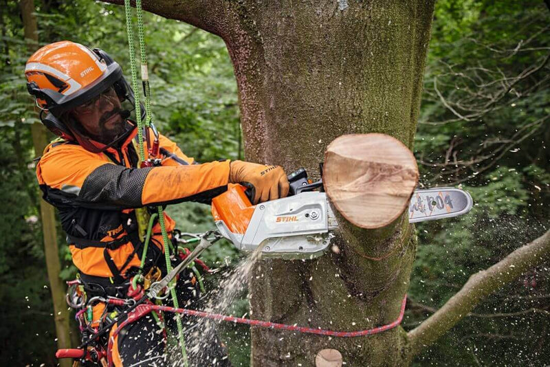 Tree worker using a chainsaw to cut wood while safely harnessed in a tree, surrounded by greenery.