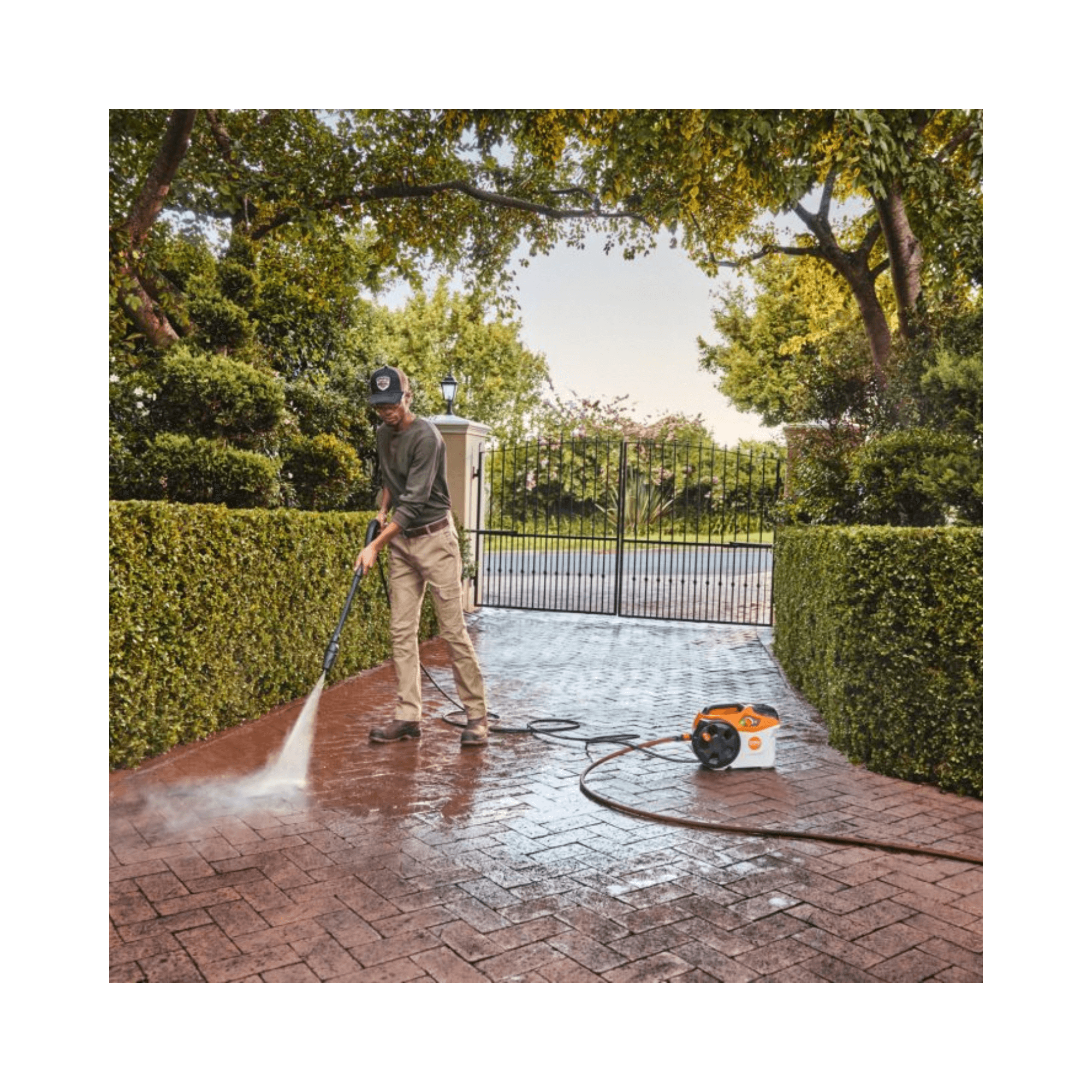 Person using REA 100 PLUS Cordless Pressure Washer on a brick driveway in a garden.