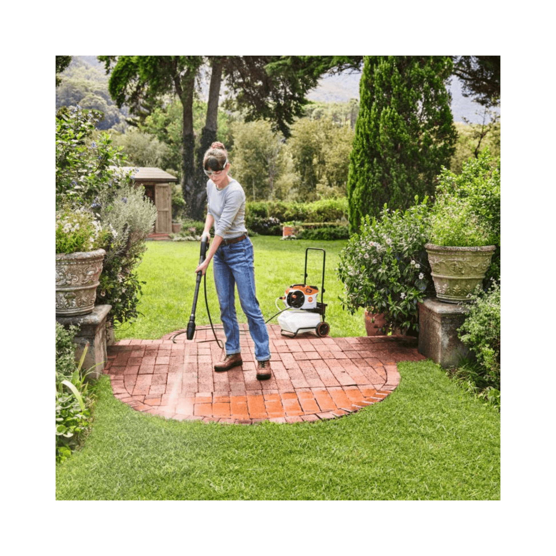 Person using a pressure washer on a garden path with trees and plants in the background