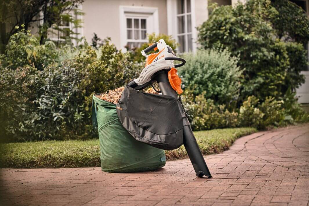 SHA 56 Cordless Leaf Vacuum Shredder beside a bag filled with leaves in a garden setting.