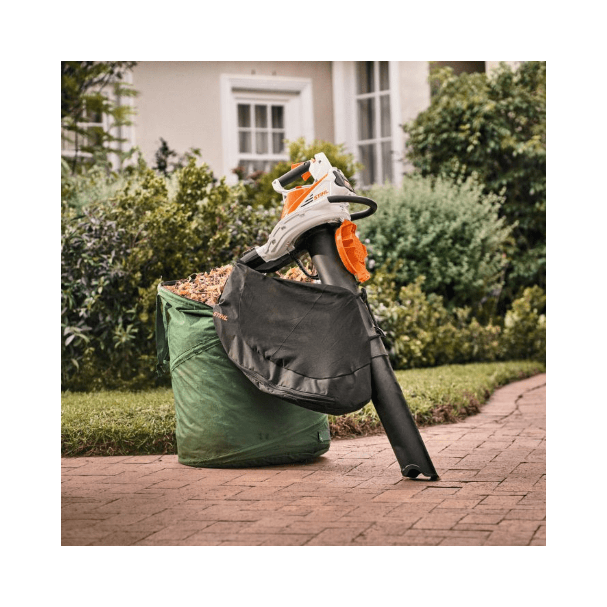 SHA 56 Cordless Leaf Vacuum Shredder with bag beside a green leaf collection bag in a garden setting.