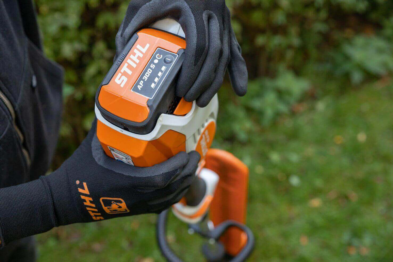 Person wearing gloves holds a STIHL battery pack for power tools in a garden setting.