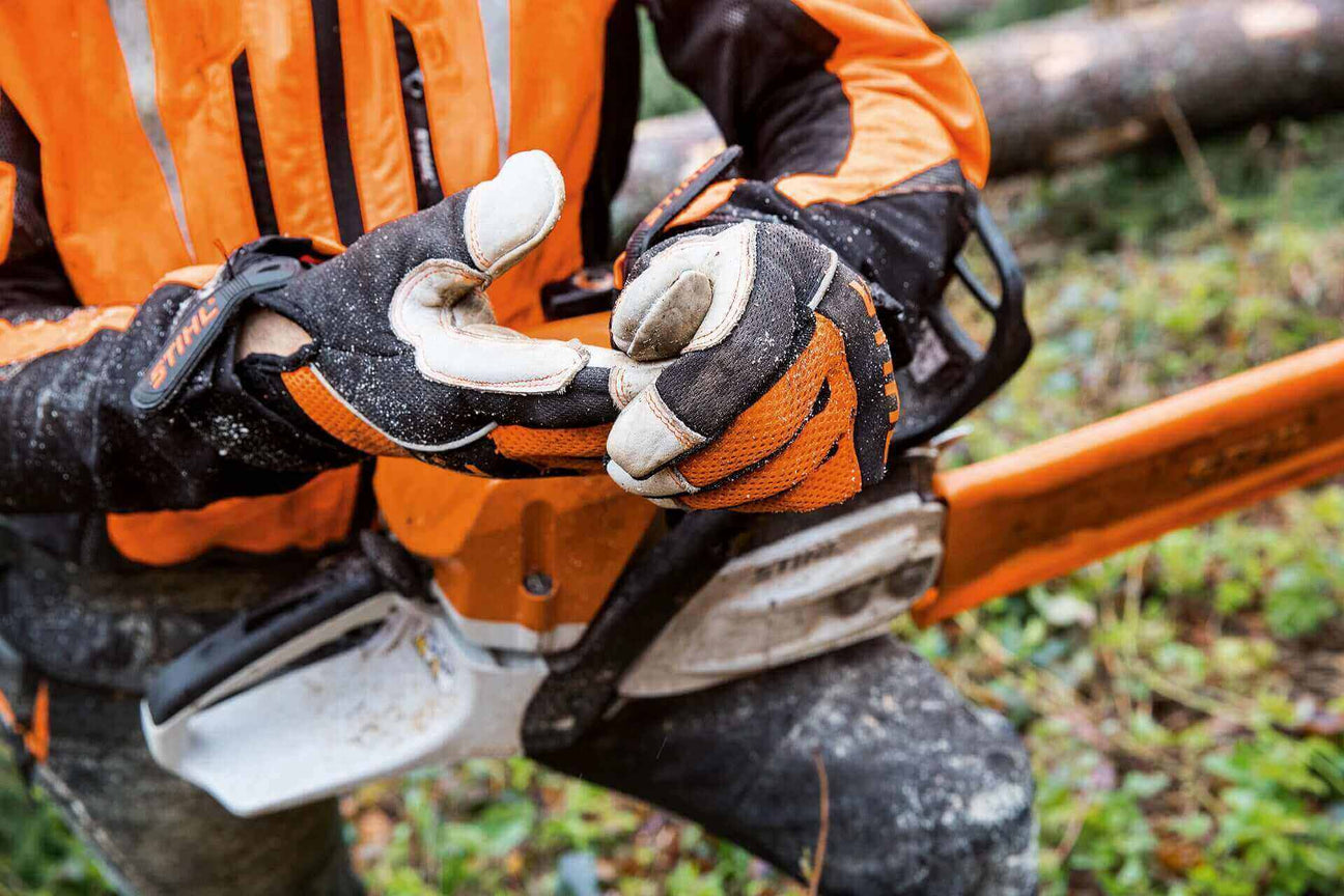 Worker in safety gear examines gloves while using a chainsaw outdoors in a forest setting.
