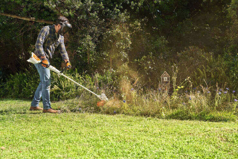 Man trimming grass with a string trimmer in a vibrant garden, wearing protective gear.