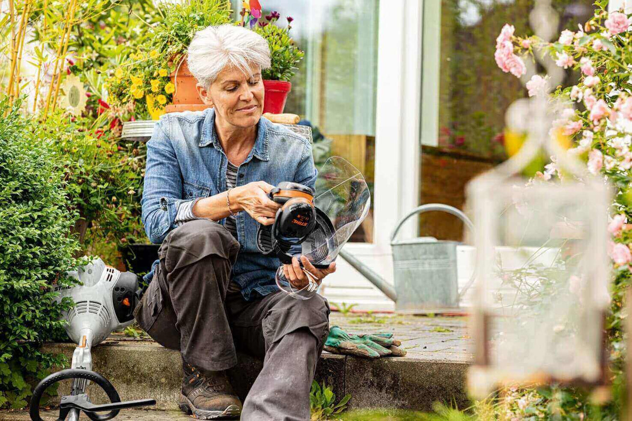 Woman gardening and using a tool in a lush outdoor space with colorful flowers.