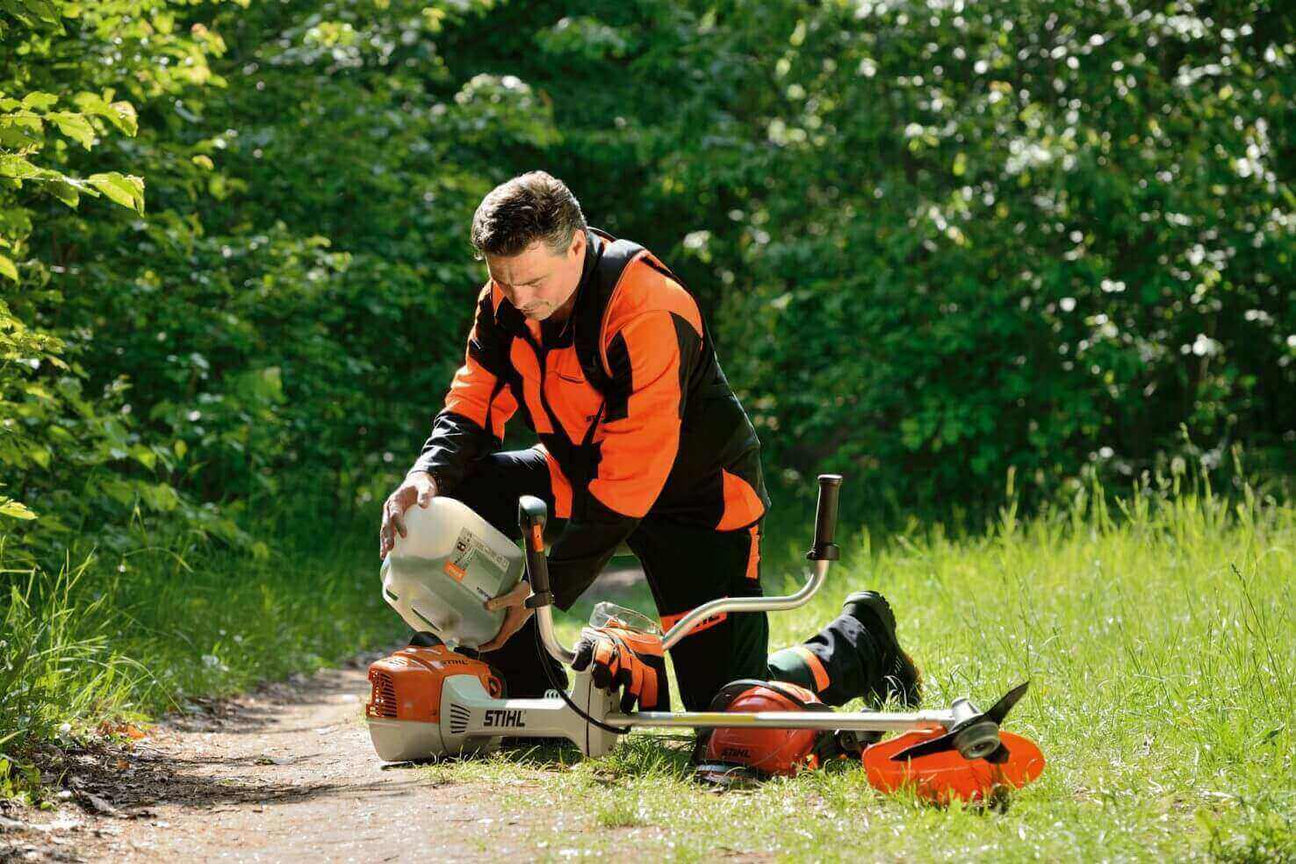 A worker in an orange and black STIHL suit kneels on a path, adjusting a trimmer on a sunny day.