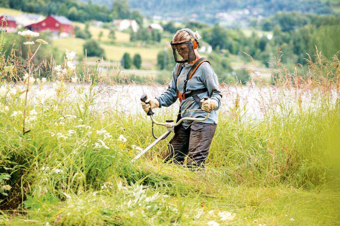 Person using a brush cutter in tall grass, wearing protective gear in a picturesque rural landscape.