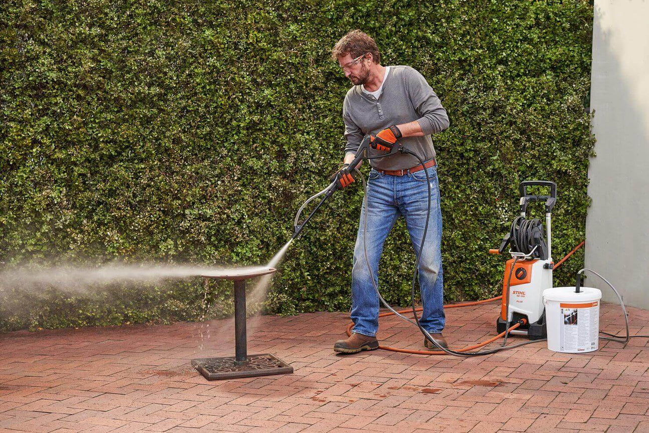 Man using a pressure washer to clean a patio table outdoors on a brick surface.