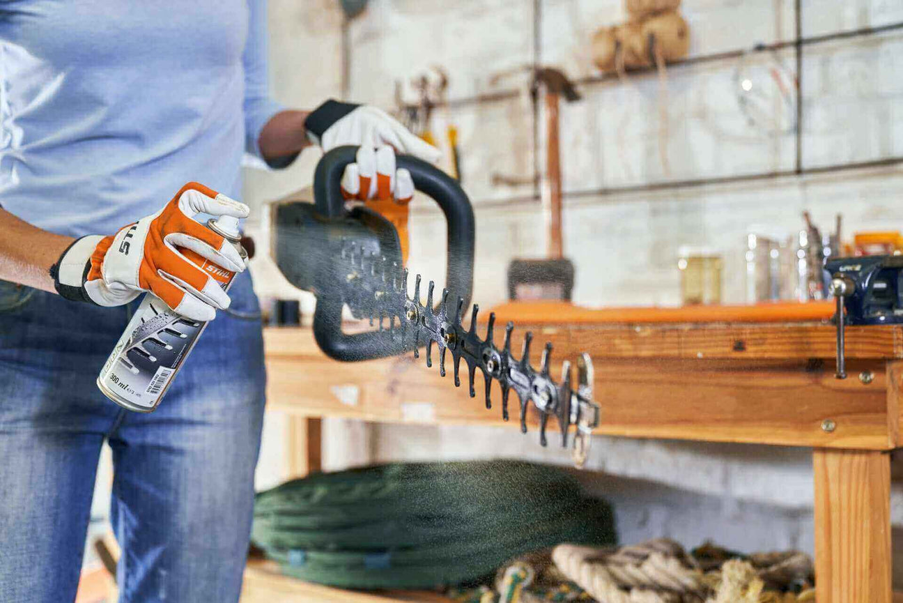 Person using a hedge trimmer while applying lubricant in a garage workshop setting.