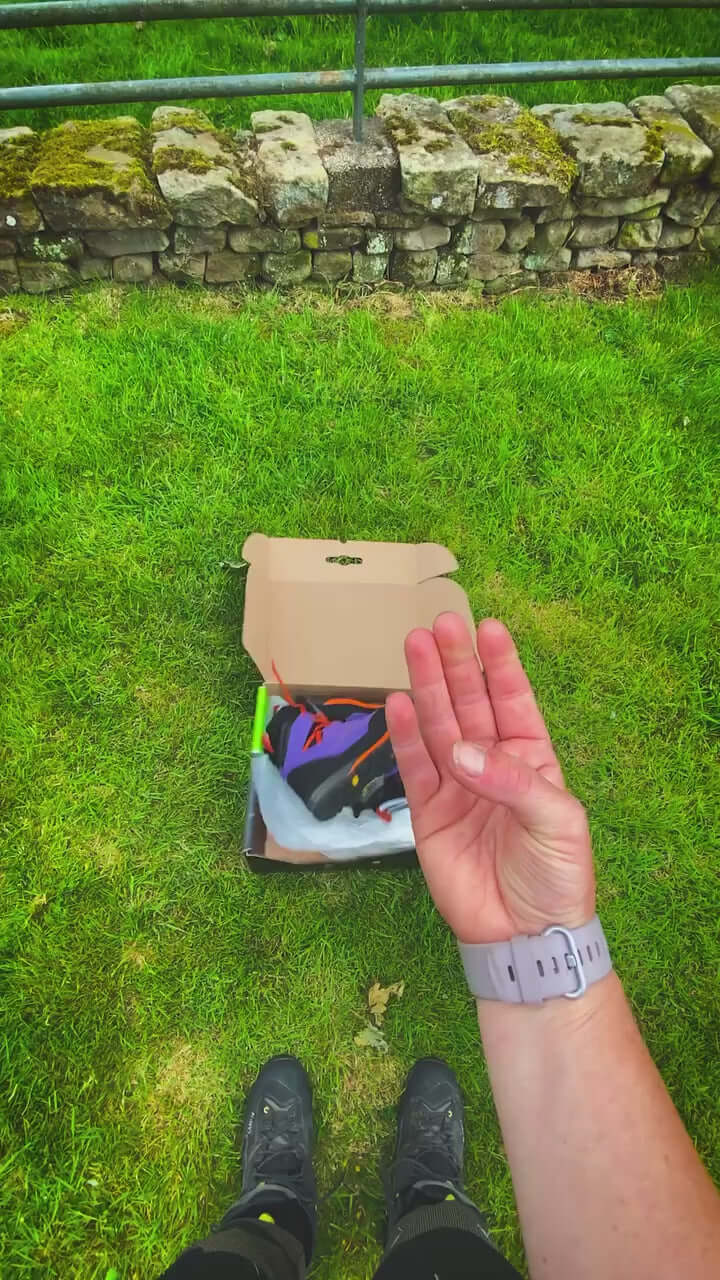 Unboxing Arbortec Kayo Purple Chainsaw Boots Class 2 on grass, showing hand gesture and footwear in a box.